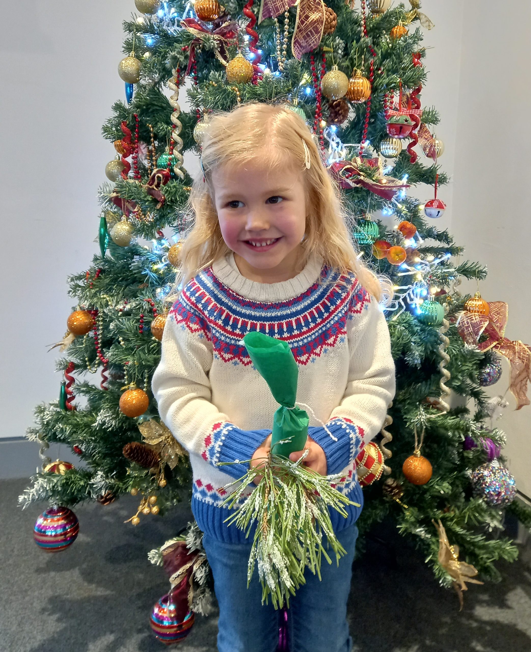 A young girl holding a Christmas decoration in front of a decorated Christmas Tree.